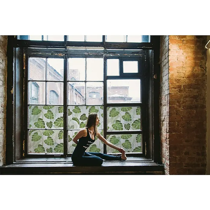 Lámina para ventana Hoja - 46 cm x 1,5 m, Verde/Transparente - Mujer practicando yoga frente a una ventana con hojas verdes como pantalla de privacidad, sentada en el alféizar con ropa deportiva.