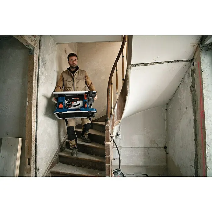 Bosch Professional AMPShare 18V Akku-Tischkreissäge GTS 18V-216 - 18 V, Ohne Akku, Durchmesser Sägeblatt: 216 mm, Ohne Sägetisch - Ein Mann trägt eine blaue Bosch-Tischkreissäge die Treppe hinunter, während er in einem unfertigen Raum arbeitet.