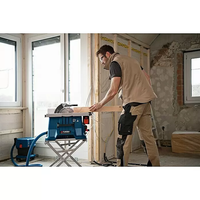 Bosch Professional AMPShare 18V Akku-Tischkreissäge GTS 18V-216 - 18 V, Ohne Akku, Durchmesser Sägeblatt: 216 mm, Ohne Sägetisch - Mann schneidet Holz mit einer blauen Tischsäge und angeschlossenem blauen Staubabsauger auf einer Baustelle.