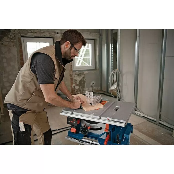 Bosch Professional AMPShare 18V Akku-Tischkreissäge GTS 18V-216 - 18 V, Ohne Akku, Durchmesser Sägeblatt: 216 mm, Ohne Sägetisch - Mann sägt Holz mit einer blauen und silbernen Akku-Tischsäge, angetrieben von einem ProCORE 18V Akku, mit Schutzausrüstung.