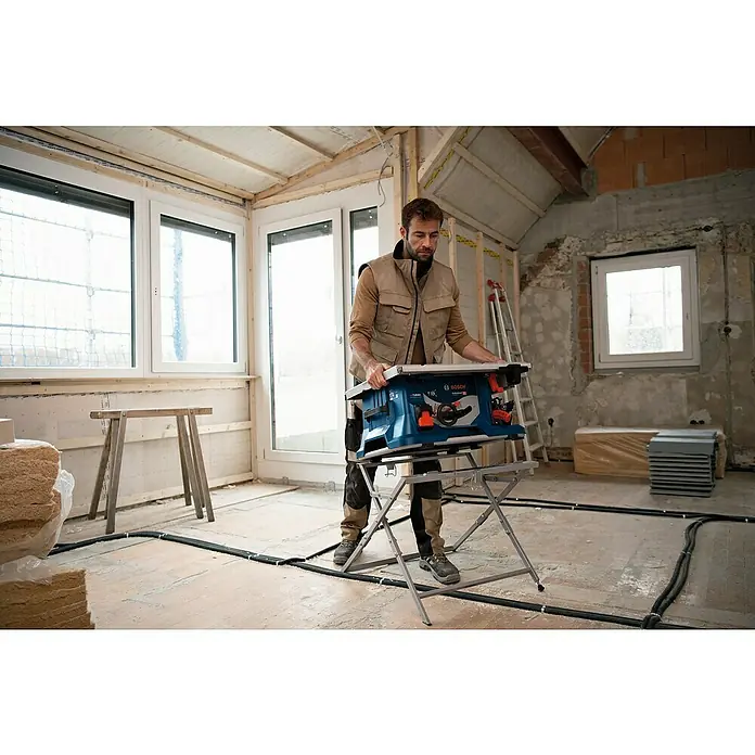 Bosch Professional AMPShare 18V Akku-Tischkreissäge GTS 18V-216 - 18 V, Ohne Akku, Durchmesser Sägeblatt: 216 mm, Ohne Sägetisch - Ein Handwerker, der in einem renovierungsbedürftigen Raum an einer blauen Bosch Tischkreissäge mit Metallgestell arbeitet.