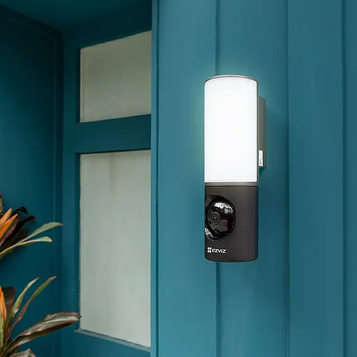 EZVIZ Überwachungskamera LC3 - Schwarz, Weiß - Sicherheitskamera mit integrierter Lampe an einer blauen Wand. Die EZVIZ-Kamera sorgt für Sicherheit und Beleuchtung im Außenbereich.