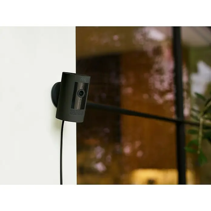 Ring Überwachungskamera außen Stick Up Cam Plug-In - Schwarz, Erfassungswinkel: 110 °, Netzbetrieben - Eine Ring-Überwachungskamera in Schwarz ist an einer weißen Wand montiert und bietet Sicherheit und Überwachung für Ihr Zuhause.
