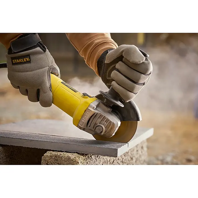 Stanley FatMax Amoladora angular eléctrica FMEG615-QS - 600 W, Diámetro disco: 115 mm, Número de revoluciones de marcha en vacío: 12.000 r.p.m. - Una amoladora angular Stanley amarilla cortando un bloque de piedra, con el operador usando guantes protectores.