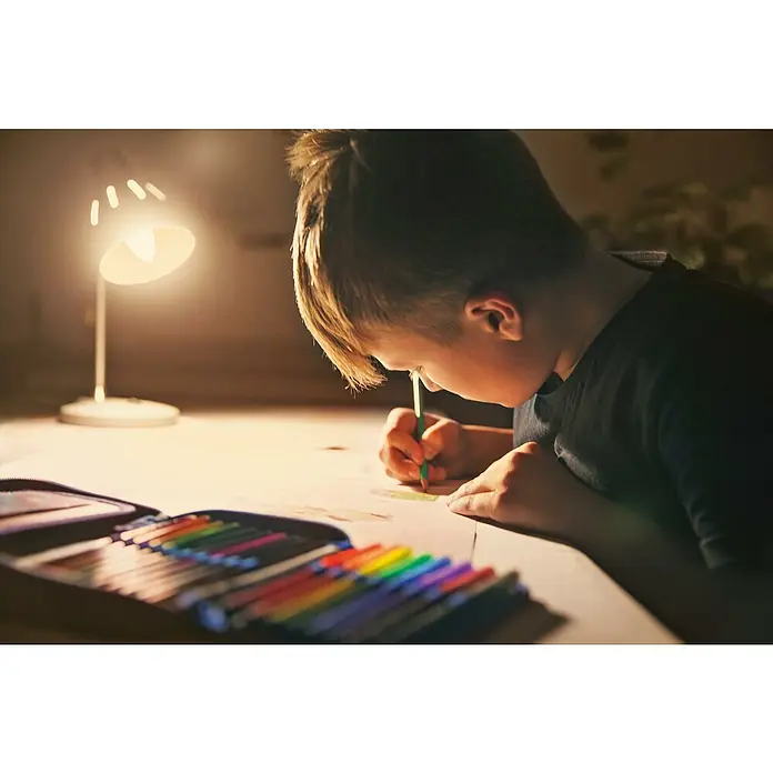 Ledvance SUN@Home Smart-LED-Lampe Smart+ WIFI - E14, Dimmbar, Neutralweiß, 425 lm, 4,9 W - Ein Kind zeichnet mit Buntstiften unter dem Licht einer Schreibtischlampe, wobei die Farben und die Konzentration des Kindes im Vordergrund stehen.