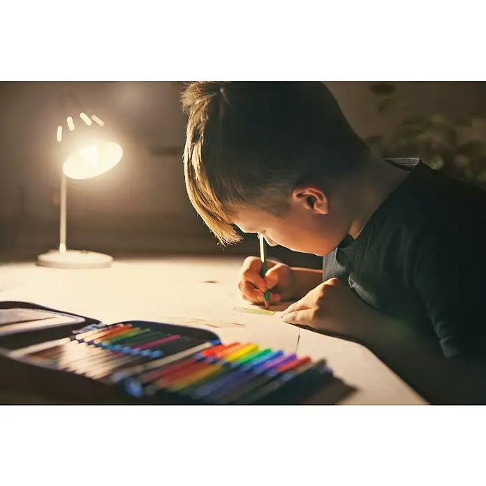 Ledvance SUN@Home Smart-LED-Lampe Smart+ WIFI - E14, Dimmbar, Neutralweiß, 425 lm, 4,9 W - Junge zeichnet mit Buntstiften unter dem Licht einer Lampe, wobei ein offenes Federmäppchen mit vielen bunten Stiften zu sehen ist.