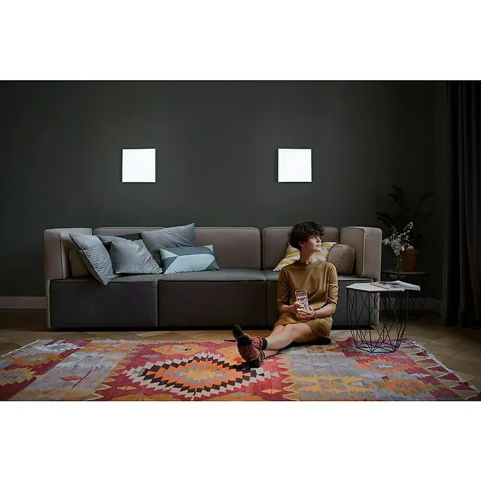 Ledvance SUN@Home LED-Panel Planon Frameless - 20 W, L x B x H: 30 x 30 x 6,1 cm, Weiß, Kaltweiß - Gemütliches Wohnzimmer mit einem Sofa, Teppich und zwei quadratischen Wandleuchten, die ein helles Licht spenden. Eine Frau sitzt auf dem Teppich.