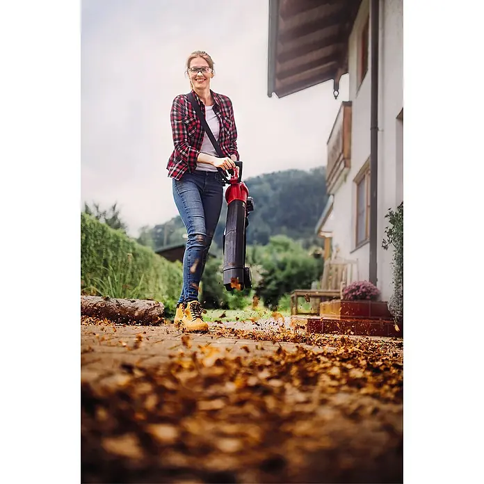 Einhell Power X-Change 18V Akku-Laubsauger GE-CL 36 Li E-Solo - Akkuspannung: 18 V, Ohne Akku, Blasgeschwindigkeit: 210 km/h - Frau benutzt einen Laubbläser, um Blätter zu entfernen. Der Laubbläser ist rot und schwarz und wird mit einem Gurt getragen.