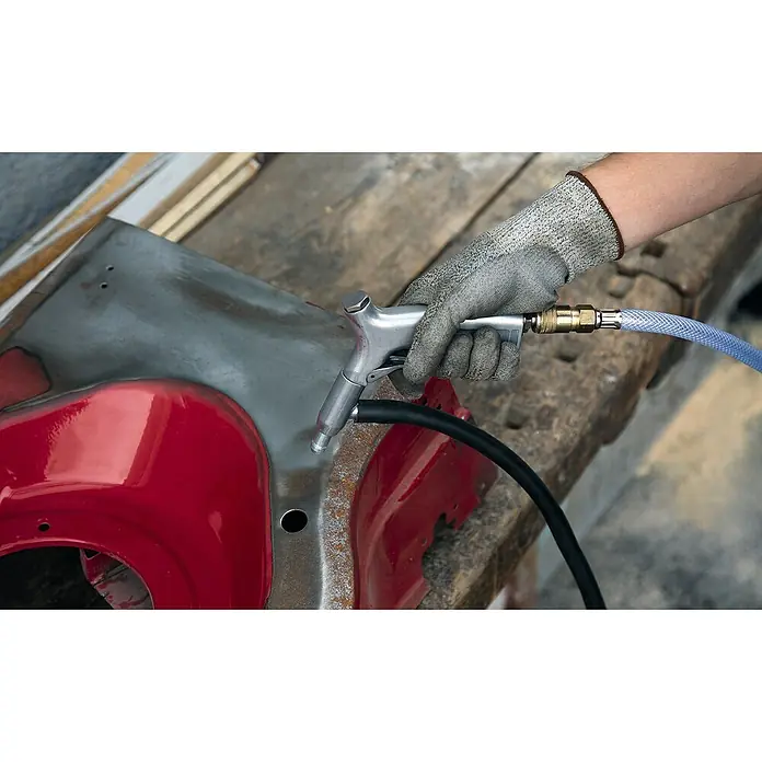 Einhell Druckluft-Saugstrahlpistole - Geeignet für: Kompressor - Detailaufnahme einer Hand mit Schutzhandschuh, die eine Sandstrahlpistole zur Metallbearbeitung an einem roten Werkstück hält.