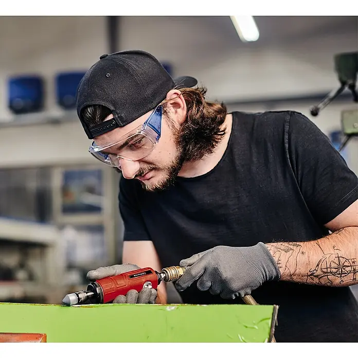 Einhell Classic Druckluft-Stabschleifer TC-PP 220 - 0 bar - 6,3 bar, 128 l/min, 22.000 U/min - Nahaufnahme eines Mannes, der mit einer roten Einhell-Schleifmaschine an einem grünen Werkstück arbeitet und dabei Schutzbrille und Handschuhe trägt.