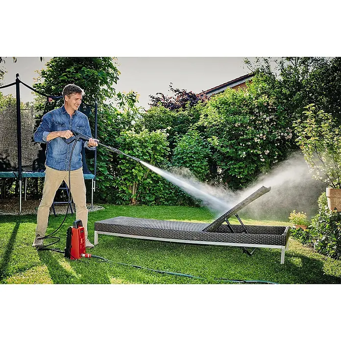 Einhell Classic Hochdruckreiniger TC-HP 90 - 1.200 W, Fördermenge: 372 l/h - Ein Mann benutzt einen Hochdruckreiniger, um eine Liege im Garten zu säubern. Der Reiniger ist rot und steht auf dem Rasen neben der Liege.