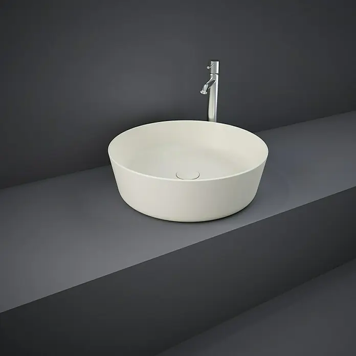 RAK Ceramics Feeling Aufsatzwaschbecken - Durchmesser: 42 cm, Ohne Spezialglasur, Hahnloch: Ohne, Ohne Überlauf, Greige, Matt - Runde Aufsatzwaschbecken aus Keramik mit chromfarbener Armatur vor einem grauen Hintergrund.