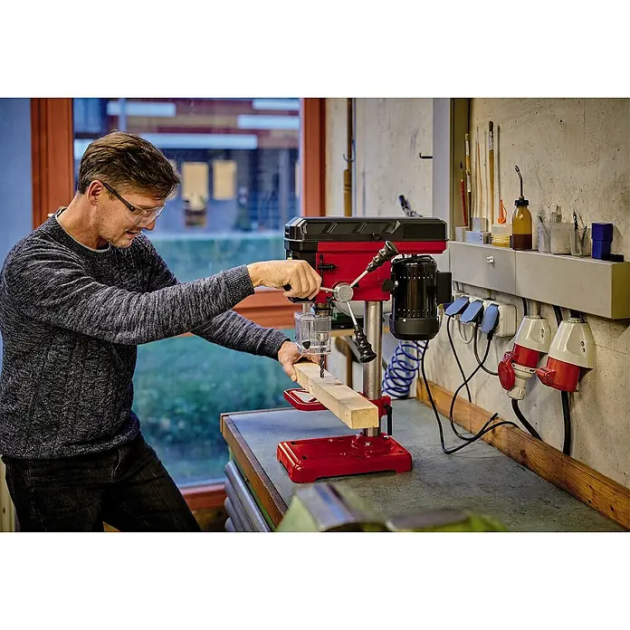 Einhell Tischbohrmaschine TC-BD 450 - 450 W, Leerlaufdrehzahl: 600 U/min - 2.650 U/min, Spannweite Bohrfutter: 1,5 mm - 13 mm - Mann bedient eine rote Tischbohrmaschine in einer Werkstatt, um präzise Löcher in ein Holzbrett zu bohren. Schutzbrille sorgt für Sicherheit.