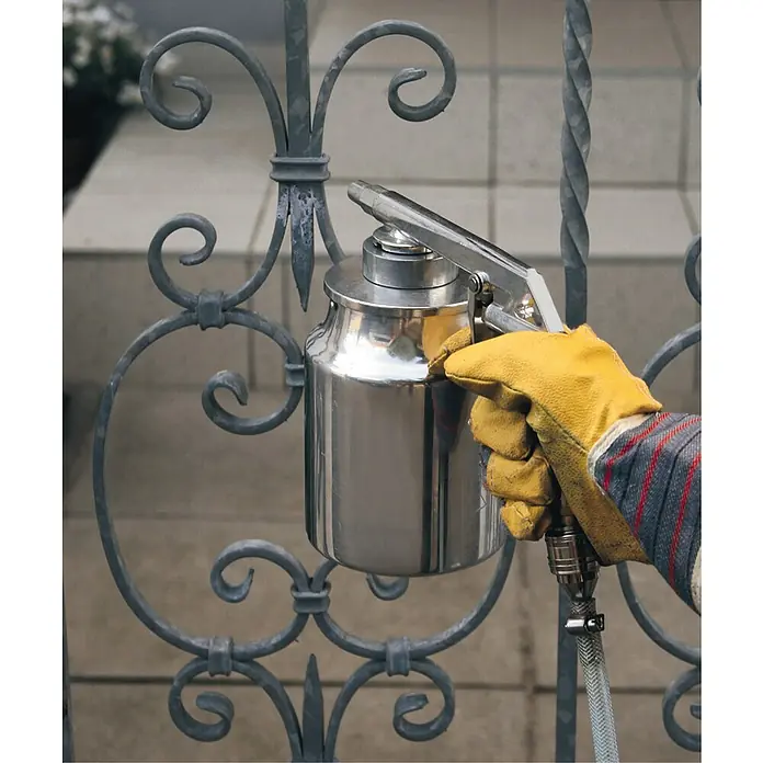 Einhell Strahlpistole mit Saugbecher - Geeignet für: Kompressor - Eine Hand mit gelbem Handschuh hält eine silberne Spritzpistole, die an einem verzierten Metalltor befestigt ist.