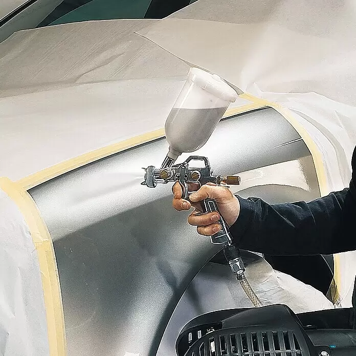 Einhell Druckluft-Farbspritzpistole mit Fließbecher - 0,6 l, 3 bar - 4 bar - Eine Hand hält eine Chrom-Lackierpistole, die ein mit Abdeckpapier versehenes silbernes Autoteil lackiert. Der transparente Becher enthält graue Farbe.