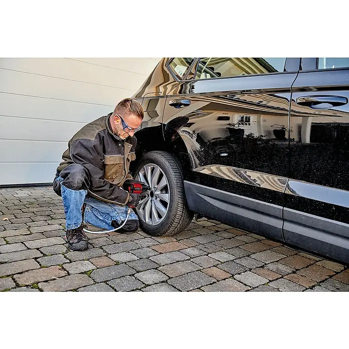 Einhell Druckluft-Set - 10 -tlg. - Ein Mann wechselt mit einem roten Einhell Akku-Schlagschrauber einen Reifen an einem schwarzen Auto vor einer Garage auf einem gepflasterten Hof.