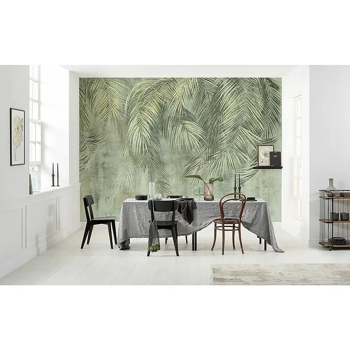 Komar Fototapete Palm FrondsB x H: 350 x 250 cm, Vlies - Elegantes Esszimmer mit einem Tisch, dekoriert mit Geschirr, und einem grünen Palmenmuster an der Wand, das eine ruhige und stilvolle Atmosphäre schafft.