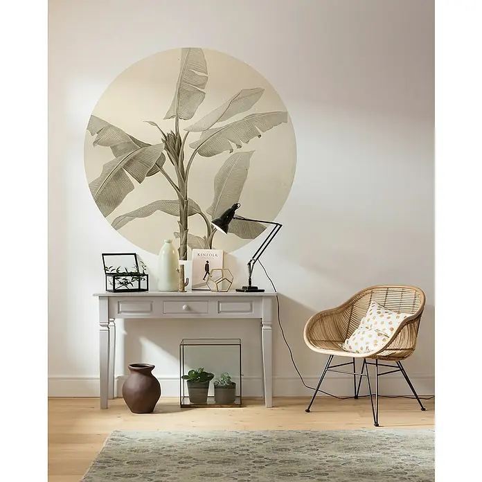 Komar Dots Fototapete rund Banana Plant - 128 cm, Selbstklebend - Wohnzimmer mit grauer Konsole, Lampe, Pflanzen und einem Rattan-Sessel mit Kissen auf einem gemusterten Teppich.