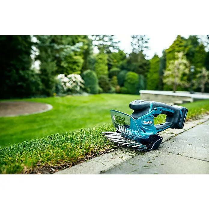 Makita Akku-Gras- & Strauchschere UM600DSMEX - 12 V, Li-Ionen, 1 Akku - Nahaufnahme einer Makita Akku-Grasschere, die auf einer Rasenkante liegt, vor einem grünen Garten.