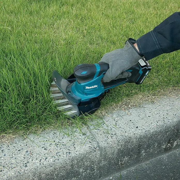 Makita Akku-Gras- & Strauchschere UM600DSMEX - 12 V, Li-Ionen, 1 Akku - Akkurasentrimmer von Makita im Einsatz, um Gras an einem Bordstein zu schneiden. Ideal für präzise Gartenarbeit und Rasenpflege.