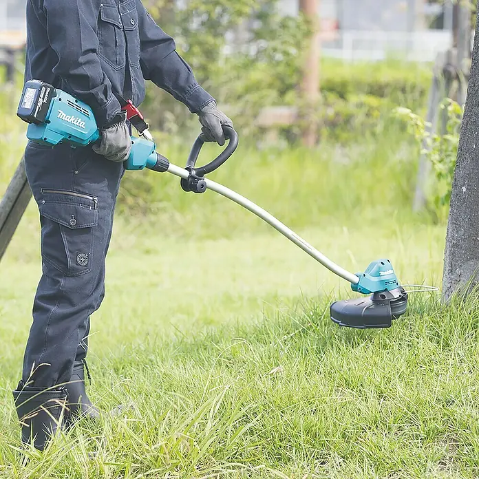 Makita LXT 18V Akku-Rasentrimmer DUR189RF - 18 V, 3 Ah, Li-Ionen, 1 Akku, Schnittbreite Faden: 300 mm - Ein Arbeiter benutzt einen Makita Akku-Rasentrimmer, um Gras um einen Baum herum zu schneiden.