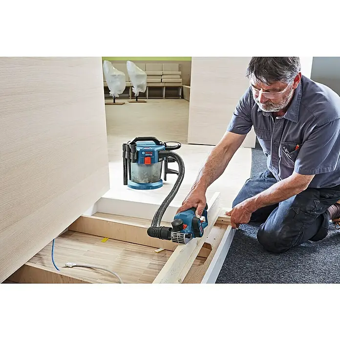 Bosch Professional Akku-Handhobel GHO 12V-20 - 12 V, Li-Ionen, Ohne Akku, Hobelbreite: 56 mm - Mann arbeitet mit einem Bosch Elektrohobel und Staubsauger, um Holz zu bearbeiten, mit Schutzbrille für Sicherheit.