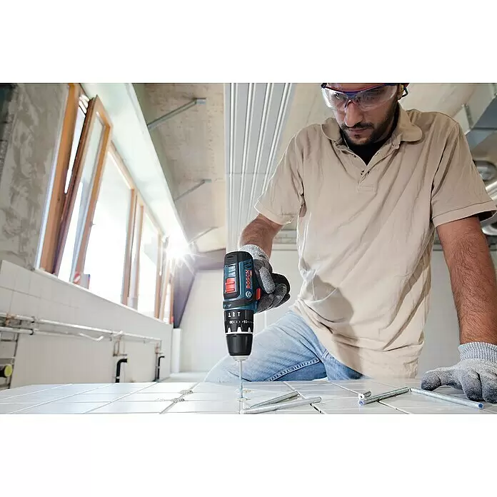 Bosch Professional Akku-Schlagbohrschrauber GSB 12V-15 - 12 V, 2 Akkus, 2 Ah, 30 Nm, 2 Akkus + Ladegerät + Bit-Set + Tasche - Ein Mann mit Schutzbrille bohrt mit einem blau-schwarzen Bosch Akkubohrer in weiße Fliesen. Gewindestangen liegen daneben.