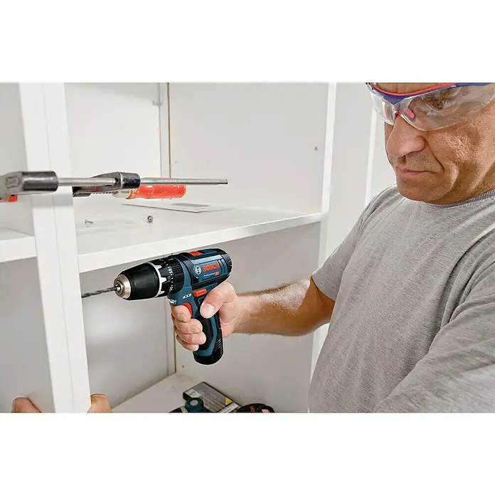 Bosch Professional Akku-Schlagbohrschrauber GSB 12V-15 - 12 V, 2 Akkus, 2 Ah, 30 Nm, 2 Akkus + Ladegerät + Bit-Set + Tasche - Ein Mann mit Schutzbrille bohrt mit einem Bosch Akku-Bohrschrauber ein Loch in ein weißes Regal. Präzise Arbeit mit hochwertigem Werkzeug.