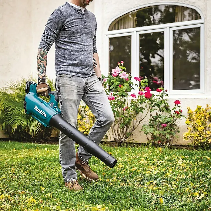 Makita LXT 18V Akku-Gebläse DUB184RF - 18 V, 1 Akku, 3 Ah, Blasgeschwindigkeit: 52,1 m/s - Ein Mann benutzt einen Makita Akku-Laubbläser im Garten, um Laub zu entfernen. Der Laubbläser ist blaugrün und schwarz.