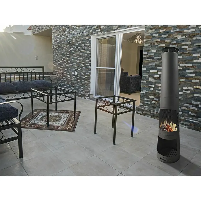 Purline Chimenea para jardín Fyn - 36 x 36 x 150 cm, Acero - Una chimenea de leña Purline gris se encuentra en un patio junto a una zona de estar, brindando calidez y ambiente.