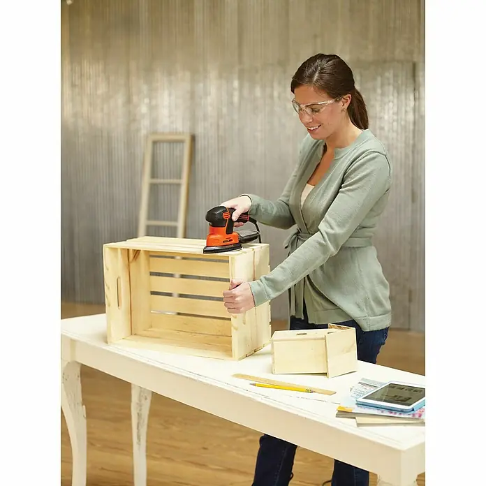 Black+Decker Elektro-Multischleifer KA2000-QS - 120 W, Leerlaufschwingzahl: 1.400 U/min, Durchmesser Schwingkreis: 2 mm - Frau schleift eine Holzkiste mit einem schwarzen und orangefarbenen Black+Decker-Schleifer auf einem weißen Tisch.