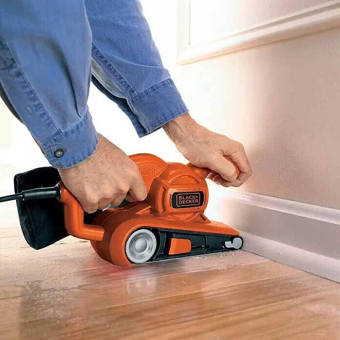 Black+Decker Elektro-Bandschleifer KA86-QS - 720 W - Oranger und schwarzer Black+Decker Bandschleifer im Einsatz, schleift einen Holzboden an einer weißen Fußleiste.