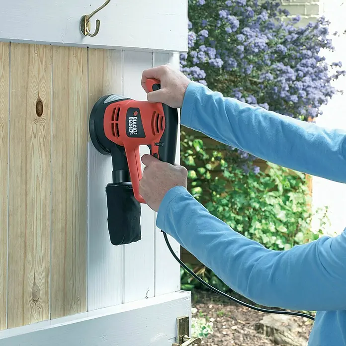 Black+Decker Exzenterschleifer KA191EK-QS - 480 W, Durchmesser Schleifteller: 125 mm - Eine Person benutzt einen Black+Decker Exzenterschleifer, um eine Holztür zu bearbeiten. Das Gerät ist orange und schwarz.