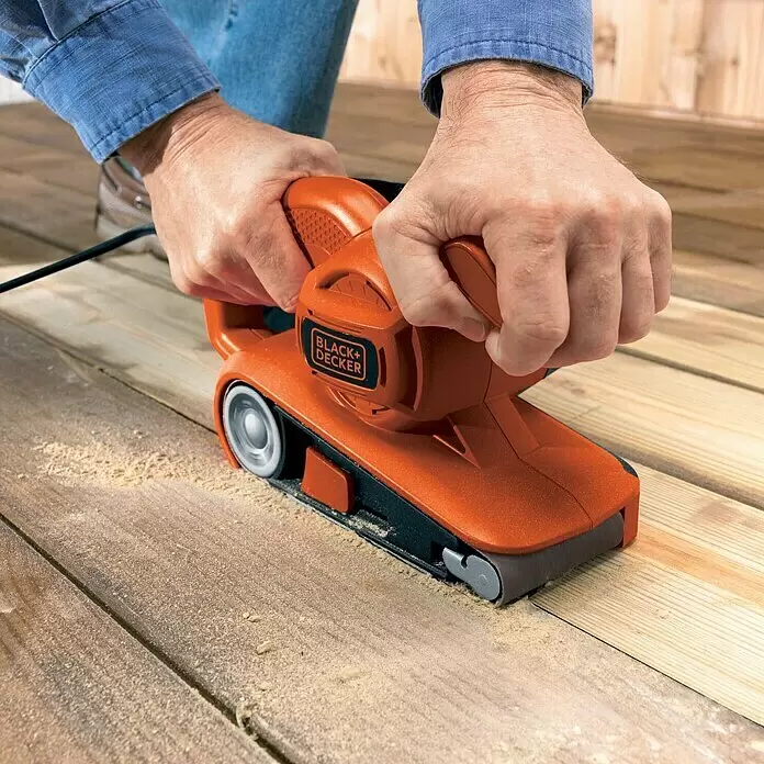Black+Decker Elektro-Bandschleifer KA86-QS - 720 W - Orange-schwarzer Black+Decker Bandschleifer glättet eine Holzterrasse und erzeugt Sägemehl.