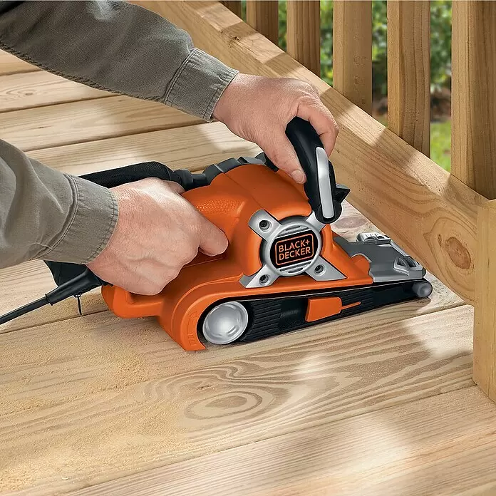 Black+Decker Elektro-Bandschleifer KA88-QS - 720 W - Person nutzt orange-schwarzen Black+Decker Bandschleifer zum Glätten eines Holzdecks, Sägespäne liegen daneben.