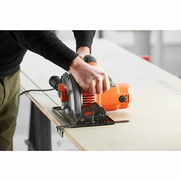 Black+Decker Handkreissäge CS1550-QS - 1.500 W, Durchmesser Sägeblatt: 190 mm, Schnitttiefe bei 90 °: 66 mm - Nahaufnahme einer Black+Decker Kreissäge beim Schneiden eines Holzbretts, bedient von einer Person mit dunkler Kleidung.