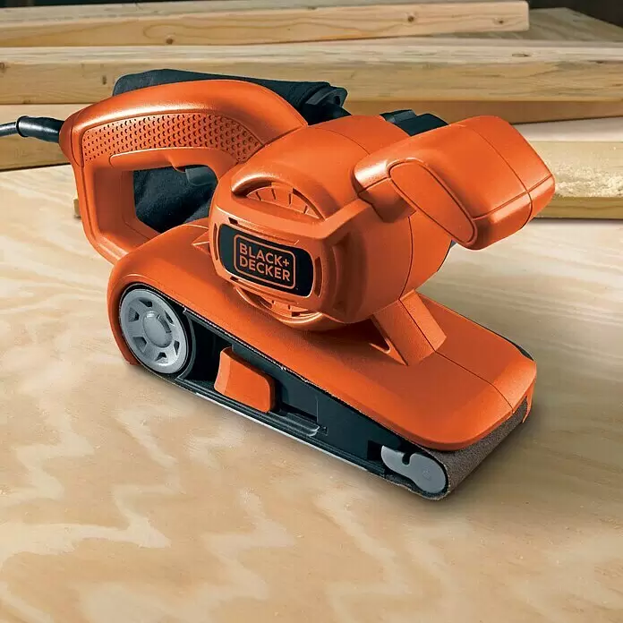 Black+Decker Elektro-Bandschleifer KA86-QS - 720 W - Orange-schwarzer Black+Decker Bandschleifer mit Staubbeutel auf hellem Holzuntergrund, mit Holzbrettern im Hintergrund.
