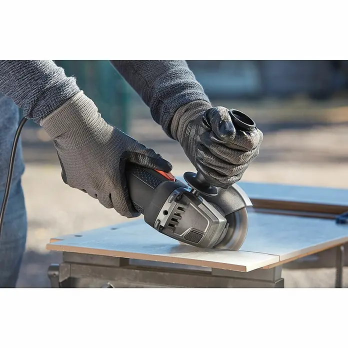 Black+Decker Elektro-Winkelschleifer BEG210-QS - 900 W, Durchmesser Scheibe: 115 mm, Leerlaufdrehzahl: 12.000 U/min - Nahaufnahme einer Person, die mit einer schwarzen Schleifmaschine eine Fliese bearbeitet und dabei Schutzhandschuhe trägt.