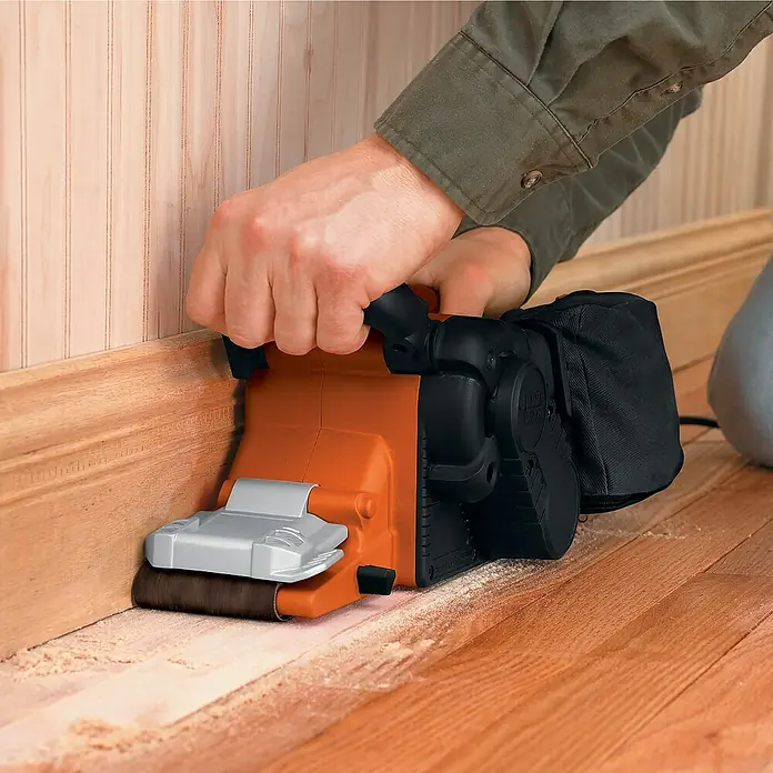 Black+Decker Elektro-Bandschleifer KA88-QS - 720 W - Nahaufnahme eines Black+Decker Kantenschleifers beim Schleifen einer Holzfußleiste und eines Holzfußbodens.