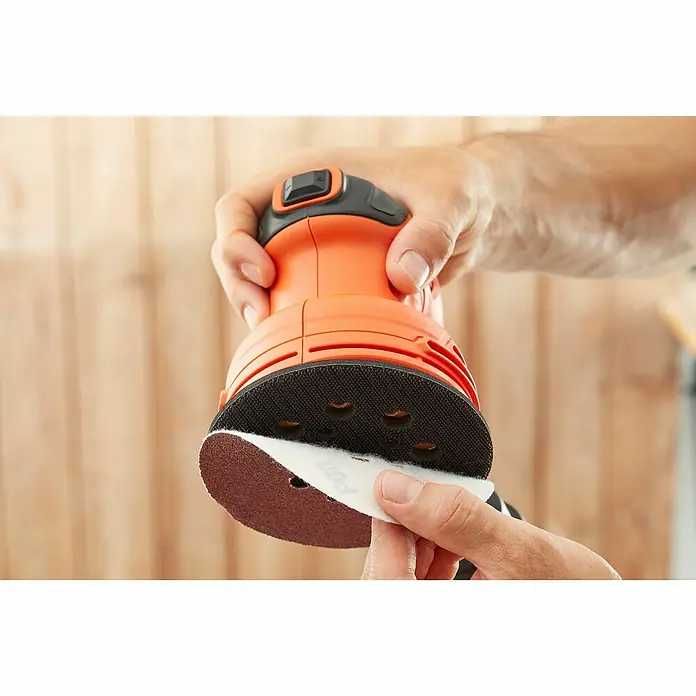 Black+Decker Elektro-Exzenterschleifer BEW210-QS - 230 W, Durchmesser Schleifteller: 125 mm - Nahaufnahme eines orangefarbenen Exzenterschleifers beim Anbringen eines Schleifpapiers mit Klettverschluss.