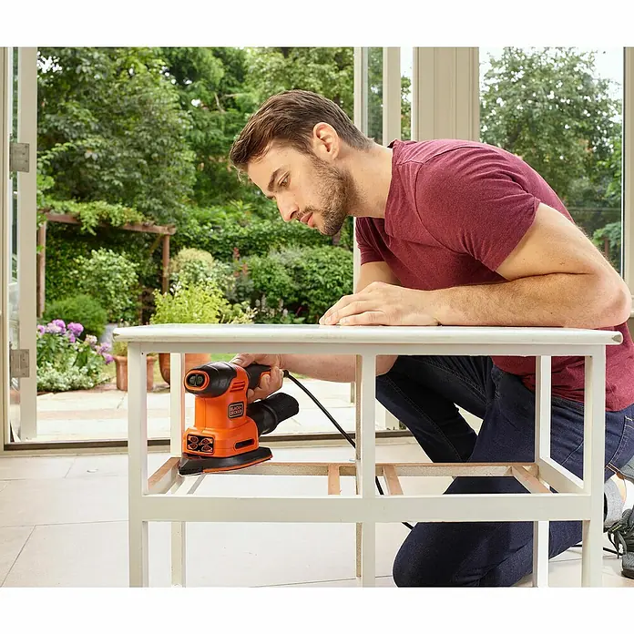 Black+Decker Elektro-Multischleifer BEW200K-QS - 200 W, Leerlaufschwingzahl: 24.000 U/min, Durchmesser Schwingkreis: 2 mm - Mann schleift mit einem Black+Decker Schleifer ein Möbelstück im Innenraum, wobei der Schleifer im Fokus steht.\n