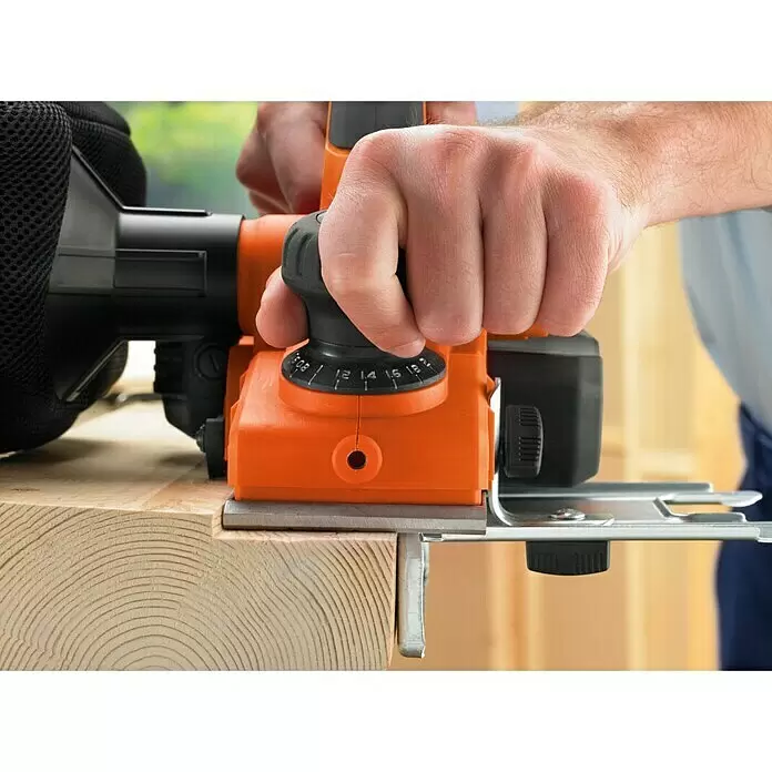 Black+Decker Elektrohobel KW750K-QS - 750 W, Hobeltiefe: 0 mm - 2 mm, Leerlaufdrehzahl: 16.500 U/min - Orangefarbener Elektrohobel mit Tiefenverstellknopf, von einer Person an einem Holzbalken verwendet.