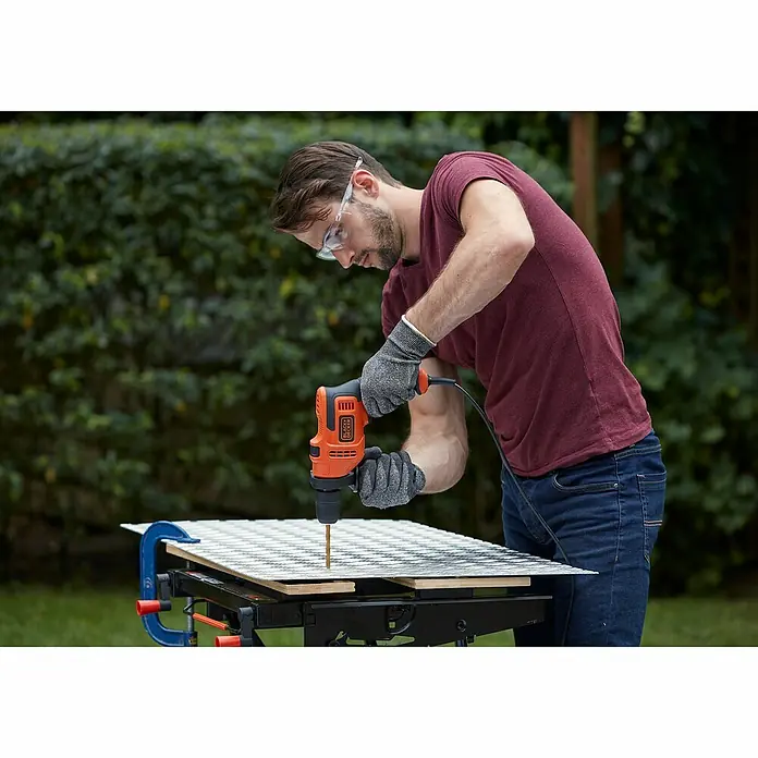 Black+Decker Schlagbohrmaschine BEH200K-QS - 500 W, Schnellspannbohrfutter - Ein Mann bohrt mit einer Black+Decker Bohrmaschine ein Loch in ein Werkstück, geschützt durch Handschuhe und Brille.