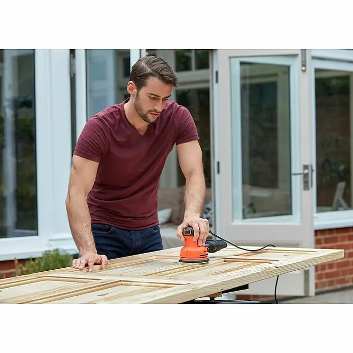 Black+Decker Elektro-Exzenterschleifer BEW210-QS - 230 W, Durchmesser Schleifteller: 125 mm - Ein Mann schleift mit einem Black+Decker Exzenterschleifer ein Holzbrett im Freien, um es für die weitere Bearbeitung vorzubereiten.