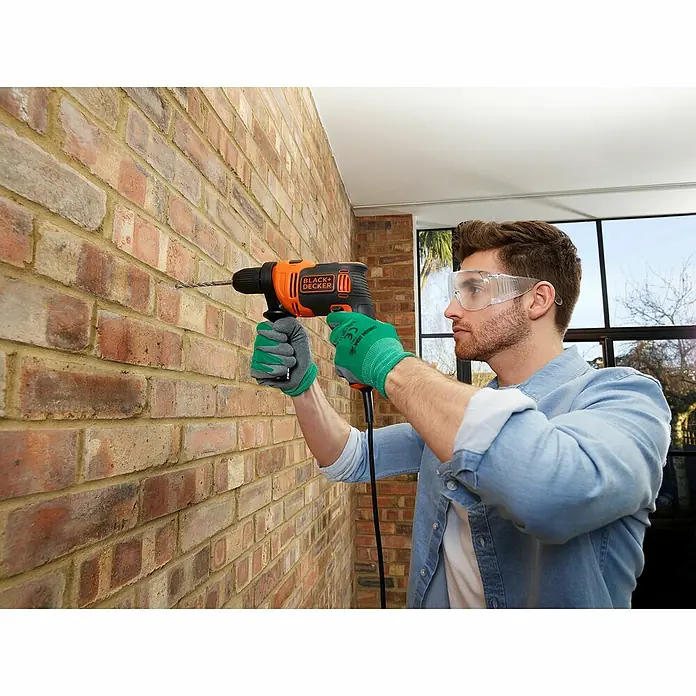 Black+Decker Schlagbohrmaschine BEH550K-QS - 550 W, Schnellspannbohrfutter - Ein Mann bohrt mit einer Black+Decker Bohrmaschine in eine Ziegelwand, geschützt durch Handschuhe und Brille.