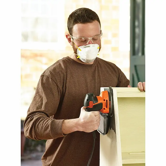 Black+Decker Elektro-Multischleifer KA2000-QS - 120 W, Leerlaufschwingzahl: 1.400 U/min, Durchmesser Schwingkreis: 2 mm - Ein Mann mit Schutzbrille und Maske schleift mit einem Black+Decker Mouse Schleifer ein Holzstück.