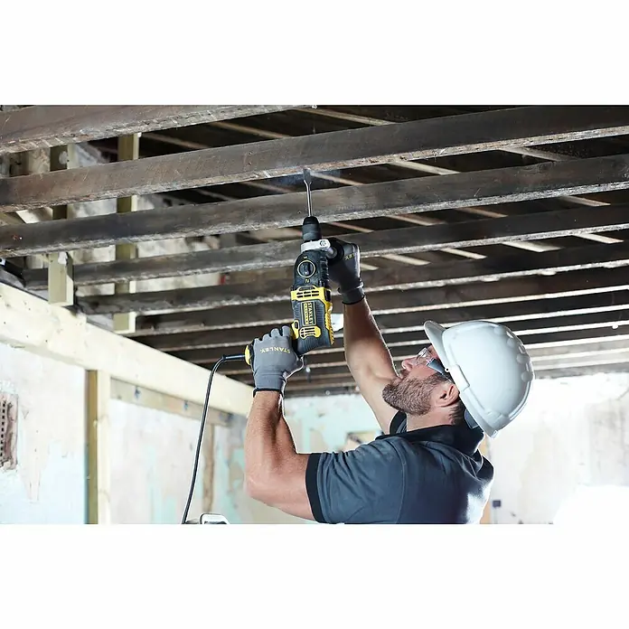 Stanley FatMax Schlagbohrmaschine FMEH850K-QS - 850 W, Schnellspannbohrfutter - Ein Mann mit Schutzhelm bohrt mit einem Stanley-Bohrhammer in eine Decke. Das Werkzeug ist gelb und schwarz und wird mit Strom betrieben.