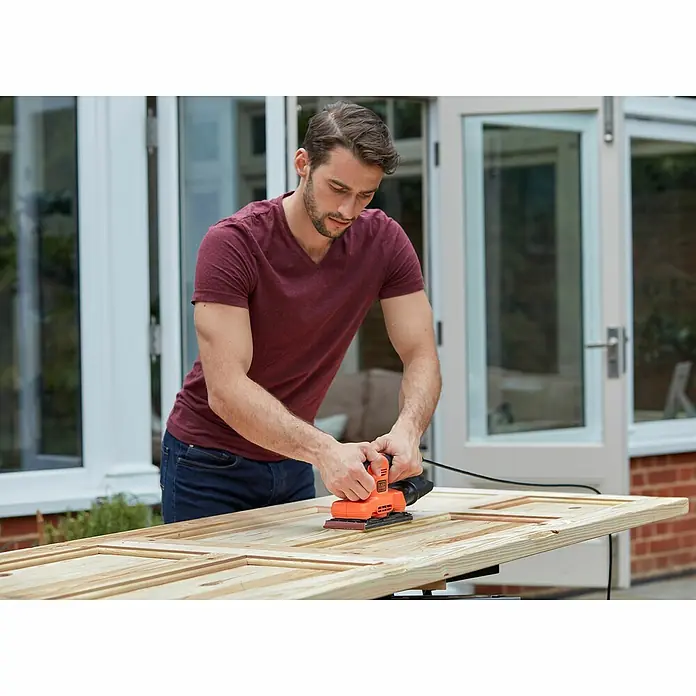Black+Decker Elektro-Schwingschleifer BEW220-QS - 150 W - Ein Mann schleift eine Holztür mit einem orangefarbenen Black+Decker-Schwingschleifer im Freien.