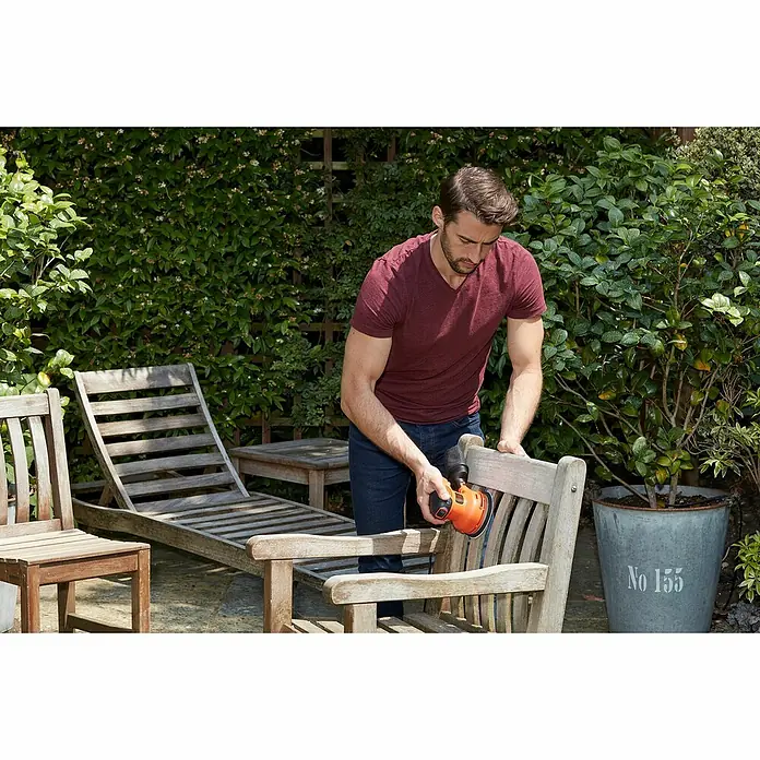 Black+Decker Elektro-Exzenterschleifer BEW210-QS - 230 W, Durchmesser Schleifteller: 125 mm - Ein Mann schleift einen Holzstuhl mit einem rot-schwarzen Exzenterschleifer im Freien, um ihn für den Sommer vorzubereiten.