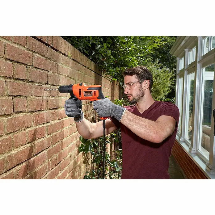 Black+Decker Schlagbohrmaschine BEH200K-QS - 500 W, Schnellspannbohrfutter - Ein Mann bohrt mit einer Black+Decker Bohrmaschine in eine Ziegelwand, geschützt durch Handschuhe und Brille, für Heimwerkerarbeiten.